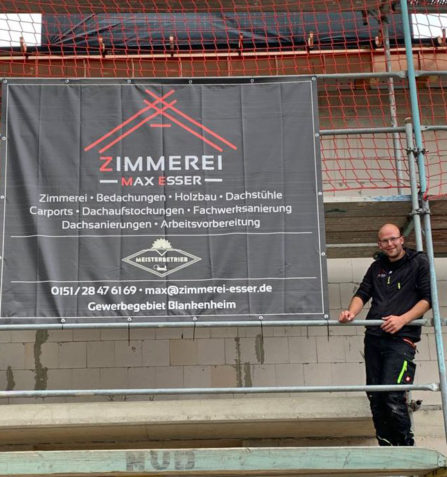 Max Esser auf einem Gerüst neben seinem Baustellenbanner der Zimmerei Max Esser
